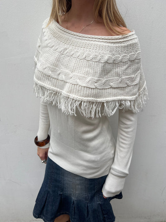 Person wearing a white knitted poncho with fringe details on a plain background