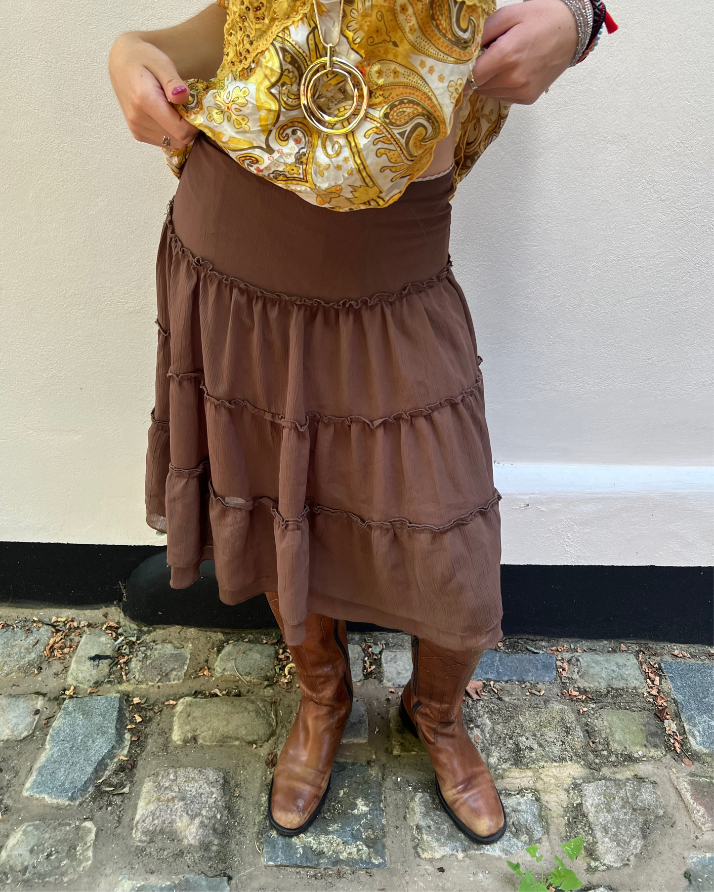 Person wearing a patterned top, brown skirt, and brown boots on a stone pavement.