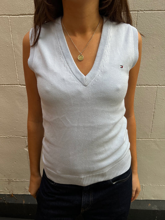 Person wearing a baby blue sleeveless sweater with a brand logo against a beige wall.