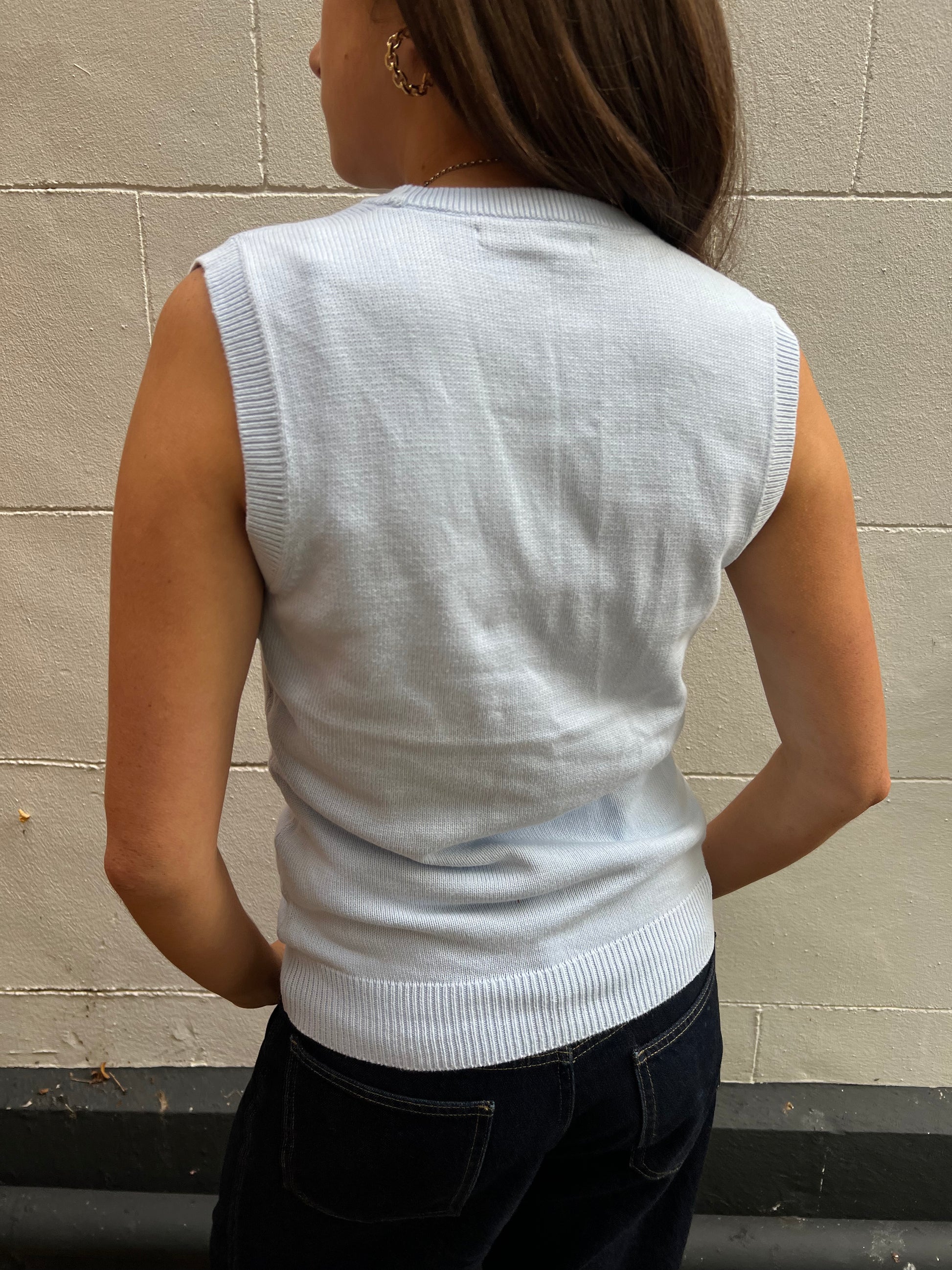 Person wearing a baby blue sleeveless sweater against a beige wall.