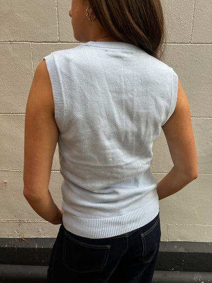 Person wearing a baby blue sleeveless sweater against a beige wall.