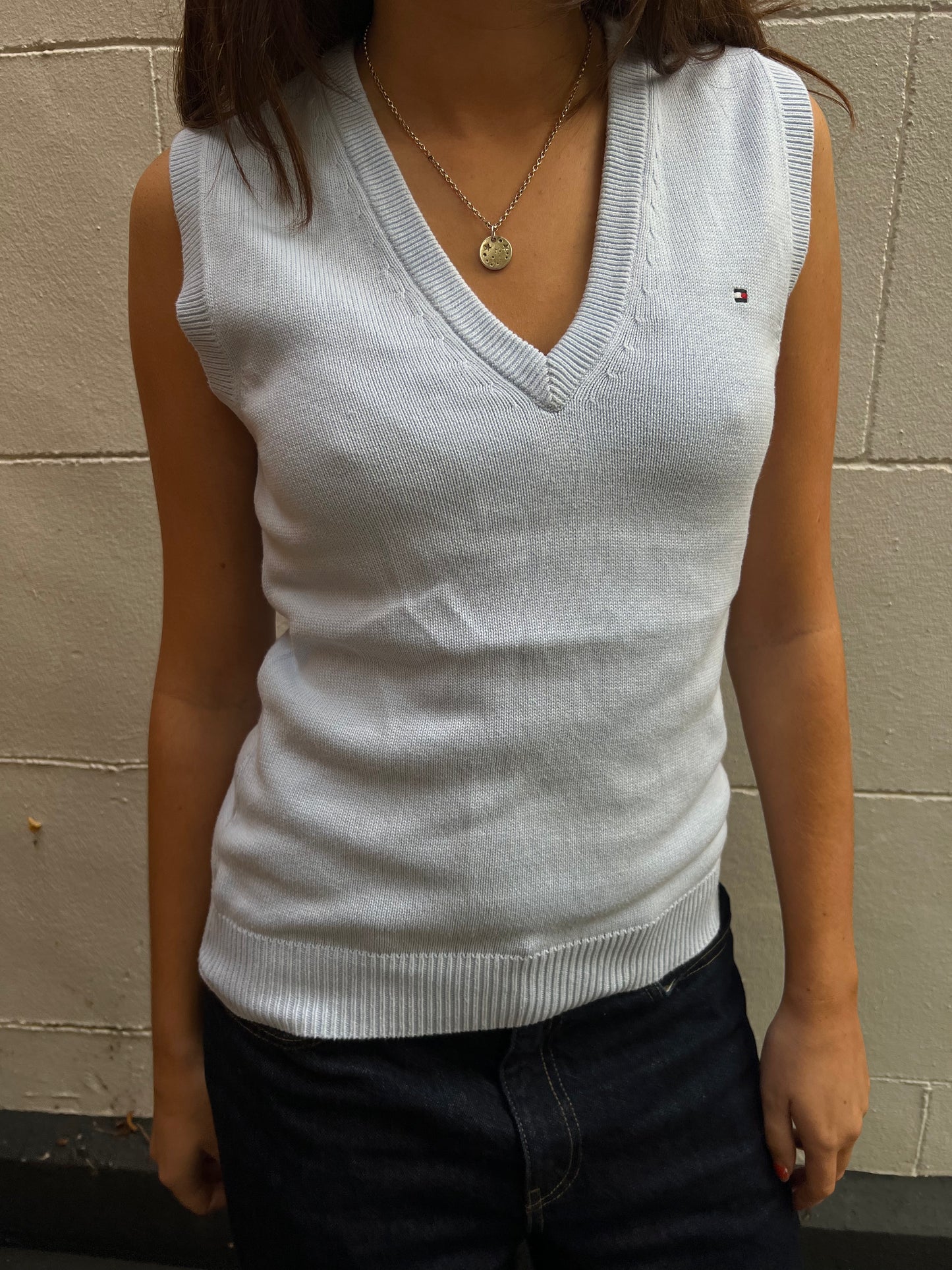 Person wearing a light blue sleeveless sweater with a brand logo against a beige wall.