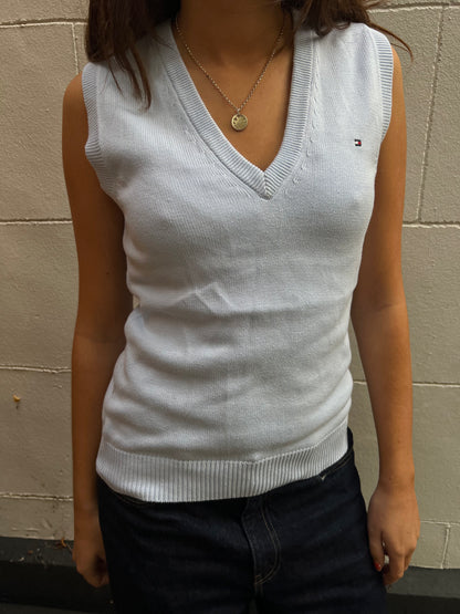 Person wearing a light blue sleeveless sweater with a brand logo against a beige wall.