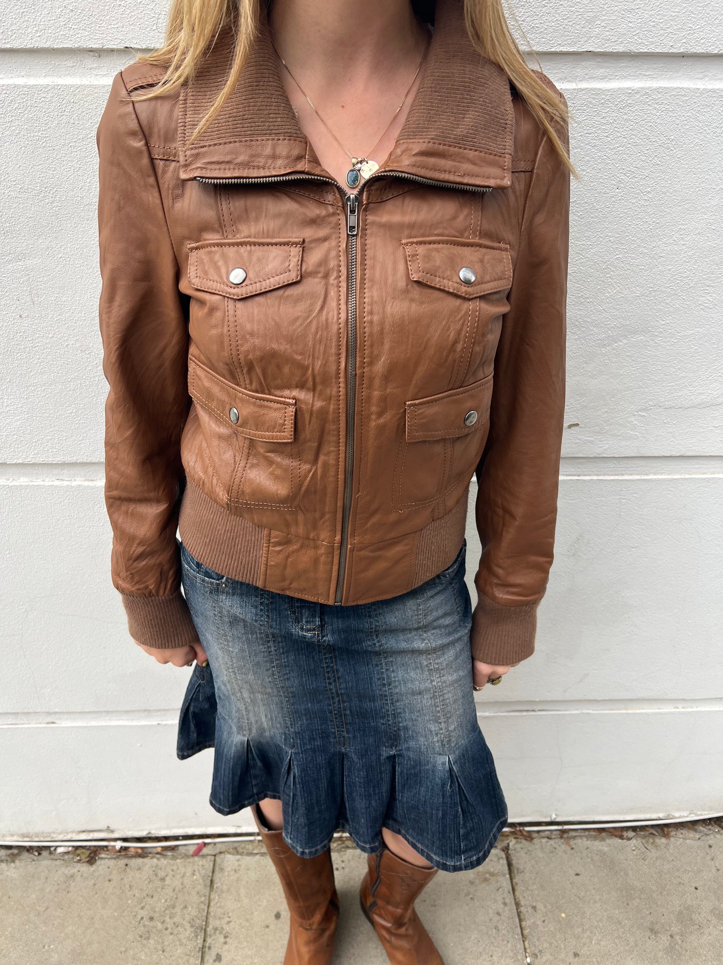 Person wearing a brown leather jacket and denim skirt against a white wall.