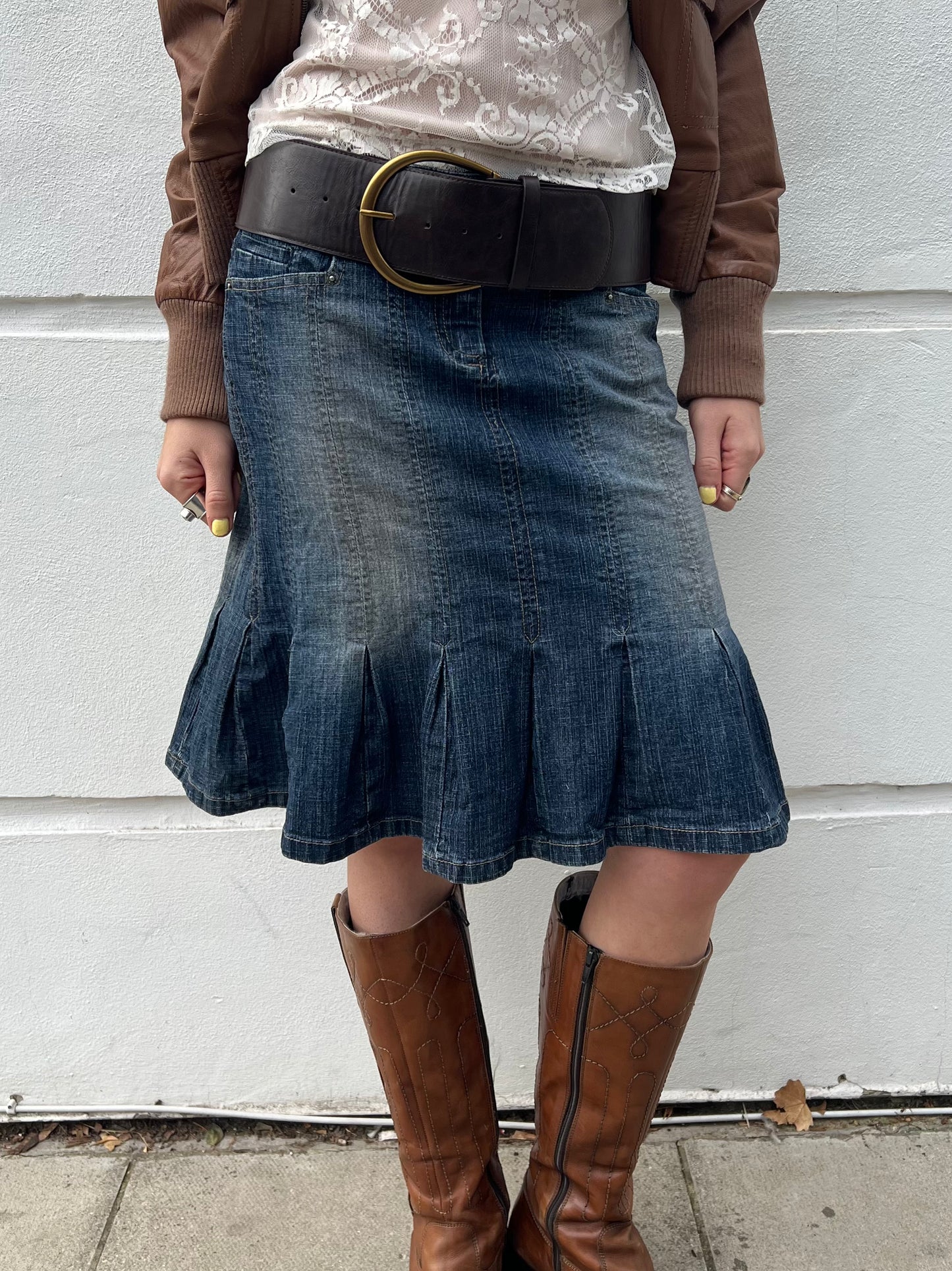 Person wearing a denim skirt and brown boots against a light gray wall.