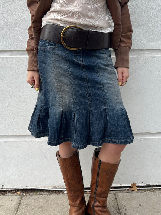 Person wearing a denim skirt and brown boots against a light gray wall.
