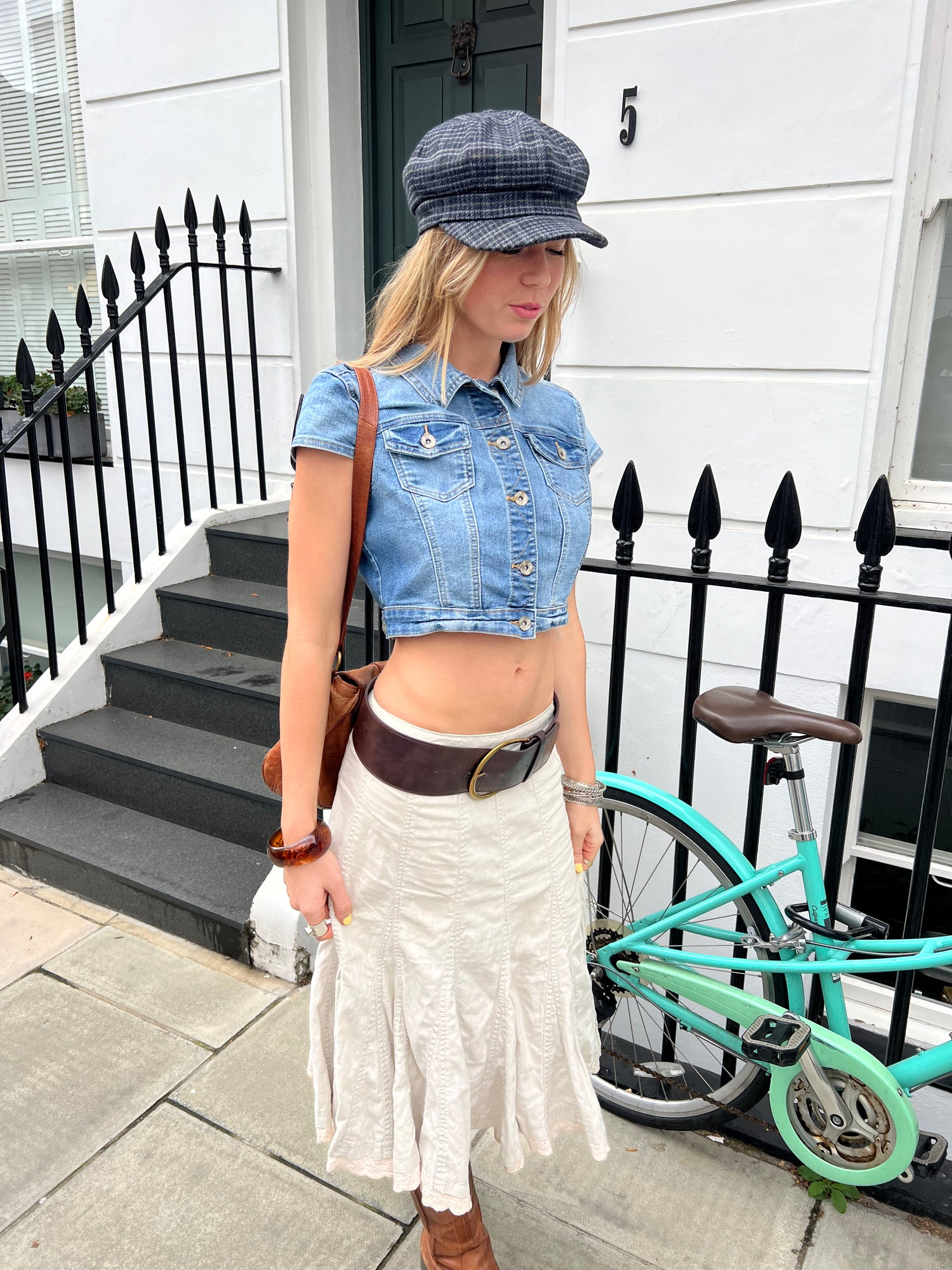 Woman in denim jacket and white skirt standing on a sidewalk with a bicycle in the background.