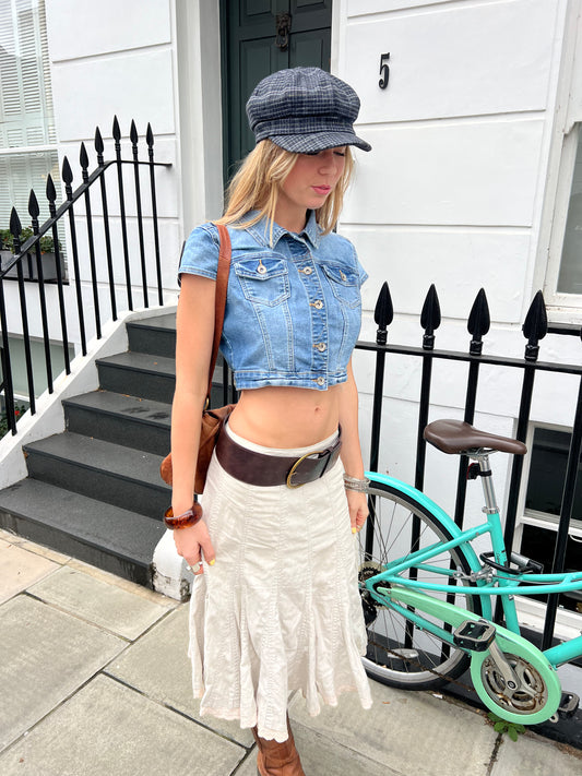 Woman in denim jacket and white skirt standing on a sidewalk with a bicycle in the background.