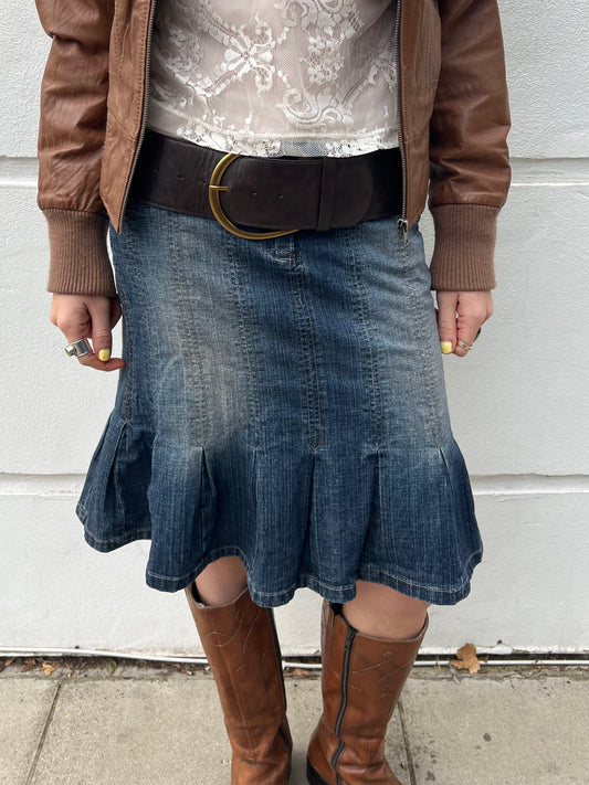 Person wearing a denim skirt, brown jacket, and knee-high boots against a white wall.