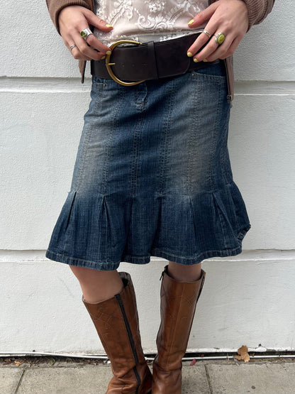 Person wearing a denim skirt with ruffled hem and brown knee-high boots against a light-colored wall.