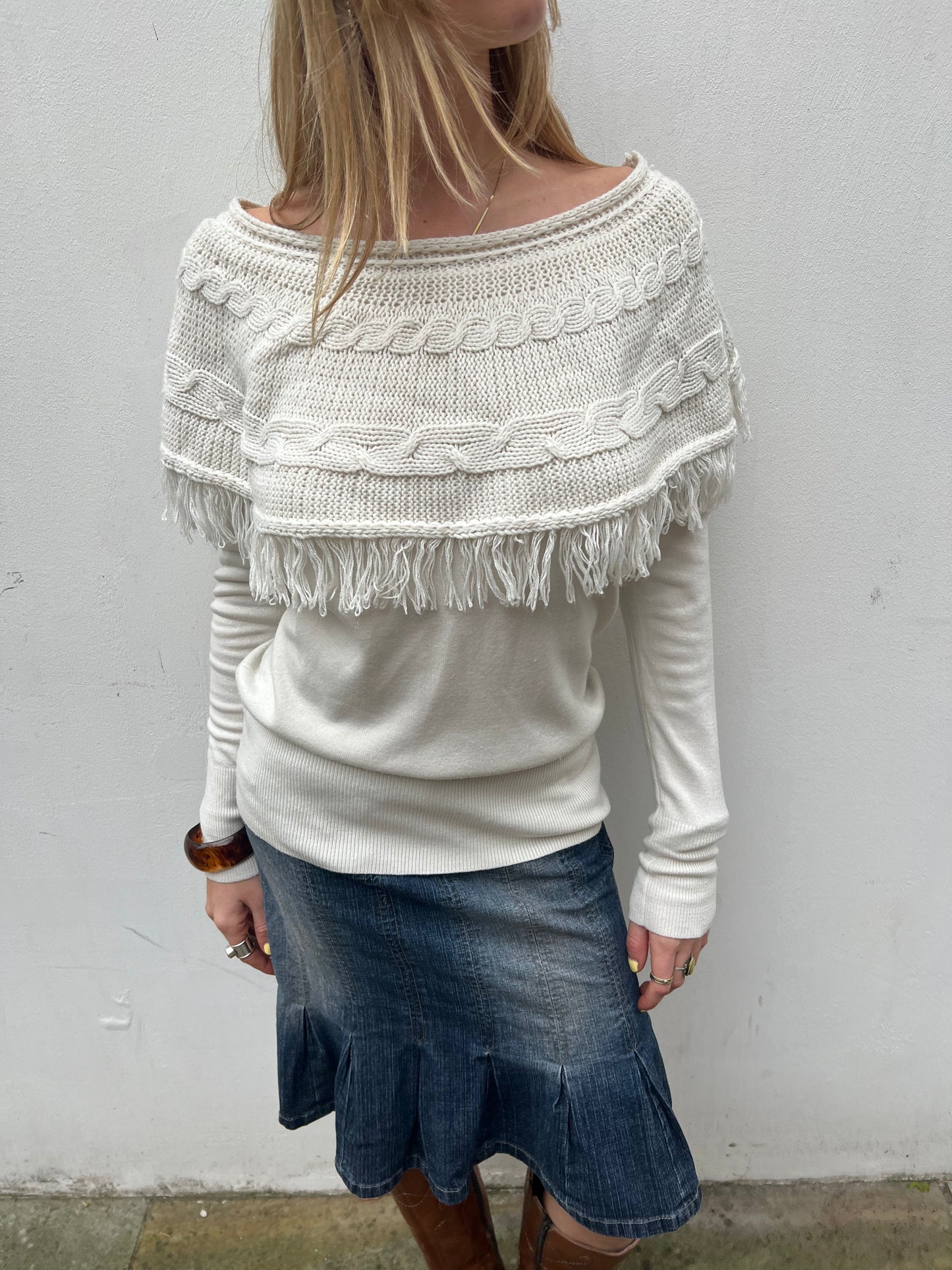 Person wearing a white sweater with fringe details and a denim skirt against a plain background