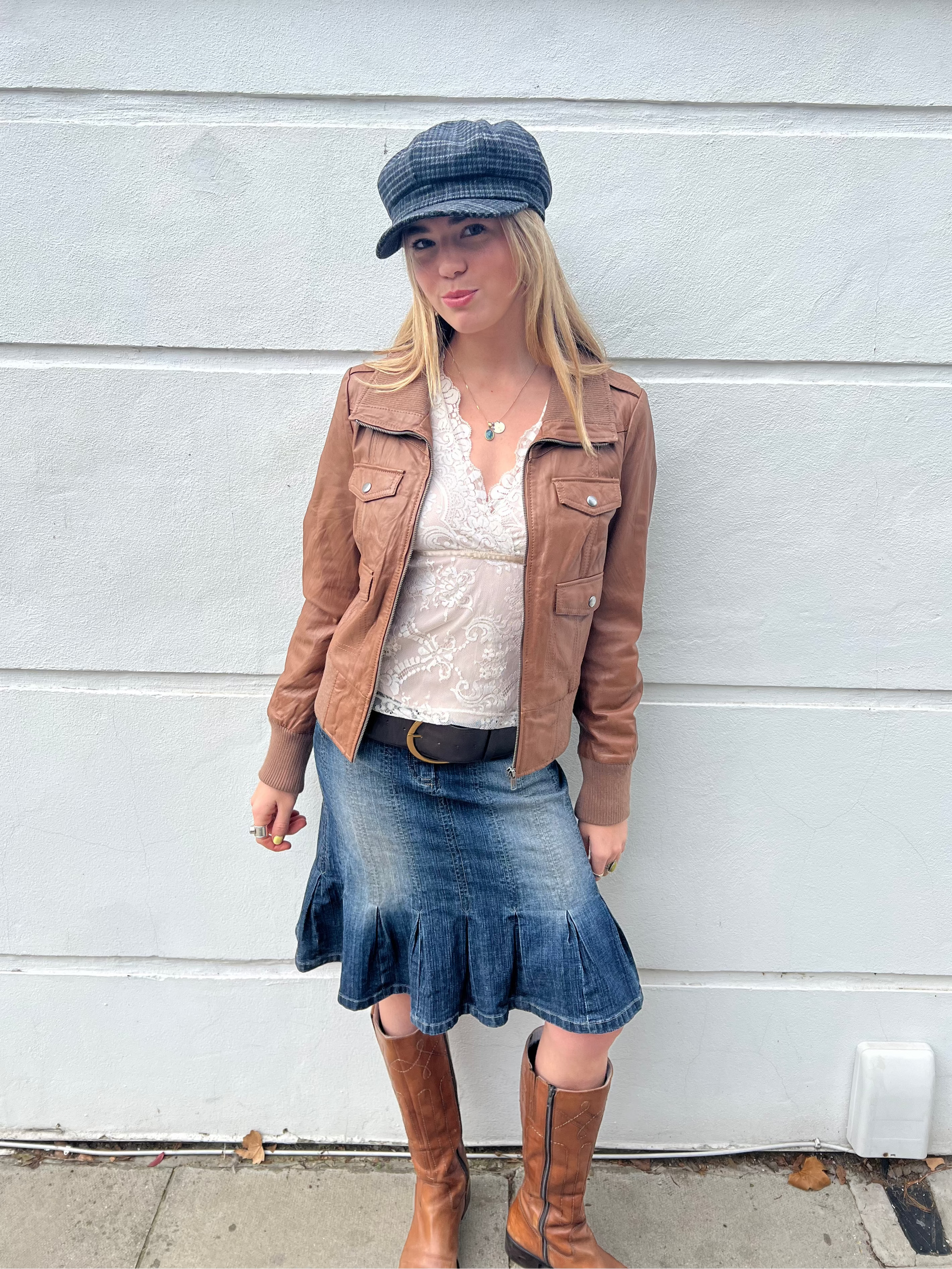 Woman wearing a brown jacket, denim skirt, and brown boots against a white wall.