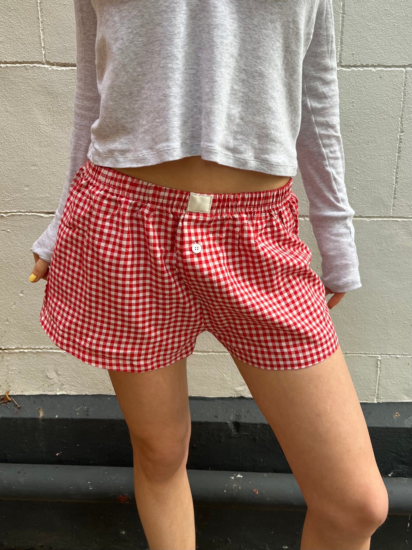 Person wearing red gingham shorts and a gray long-sleeve shirt against a gray wall.