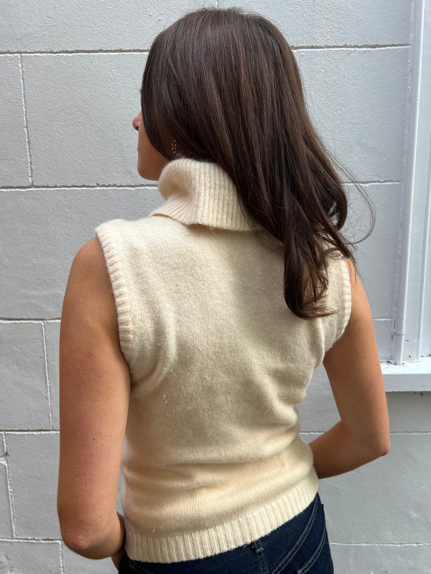 Person wearing a cream-colored sleeveless sweater against a gray tiled wall.
