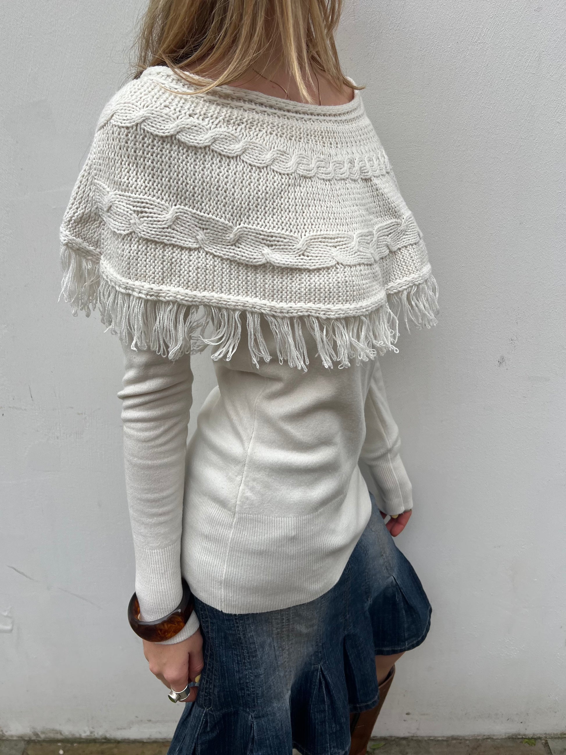 Person wearing a white knitted poncho with fringe details against a plain background