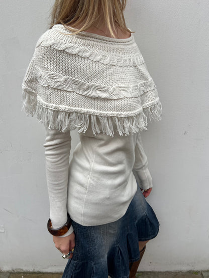 Person wearing a white knitted poncho with fringe details against a plain background
