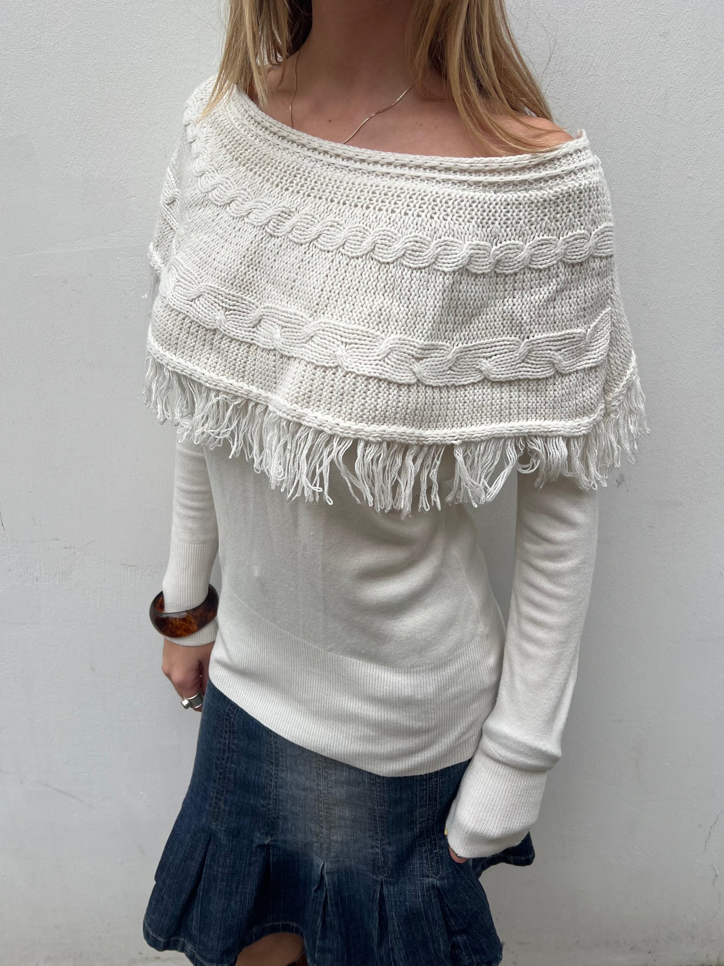 Person wearing a white knitted poncho with fringe details on a plain background