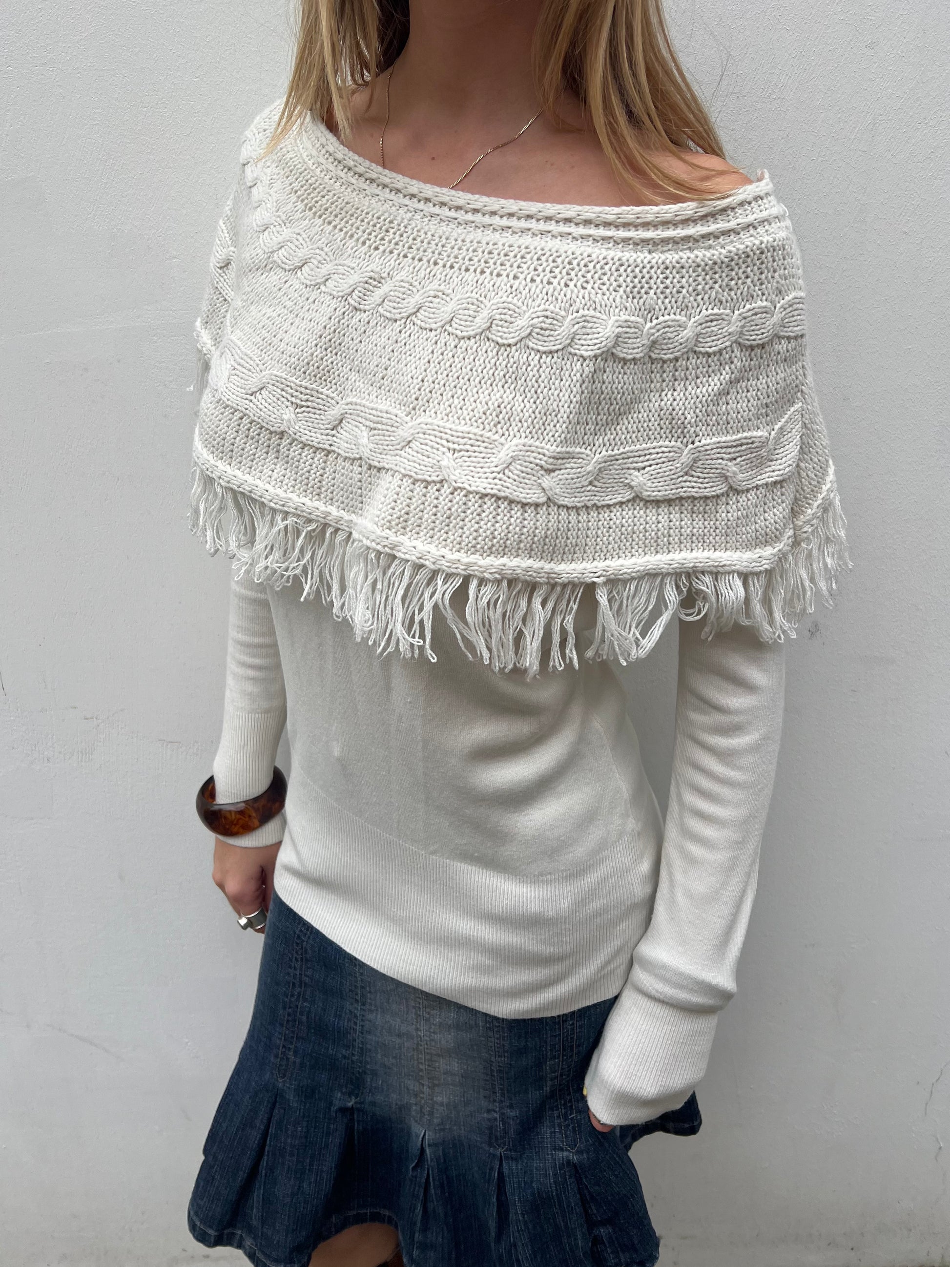 Person wearing a white knitted poncho with fringe details on a plain background
