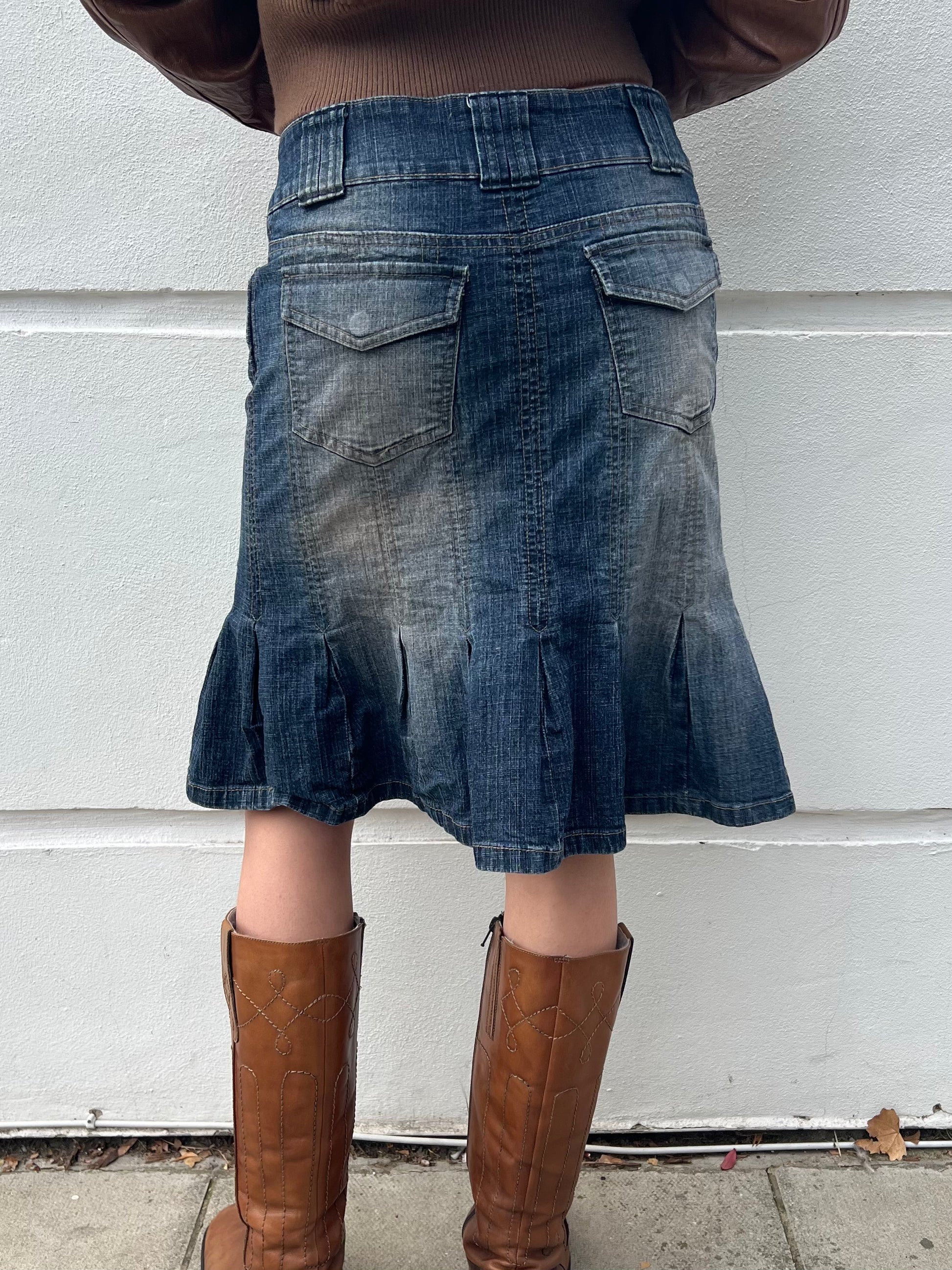 Person wearing a blue denim skirt and brown boots against a white wall.