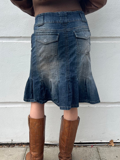 Person wearing a blue denim skirt and brown boots against a white wall.
