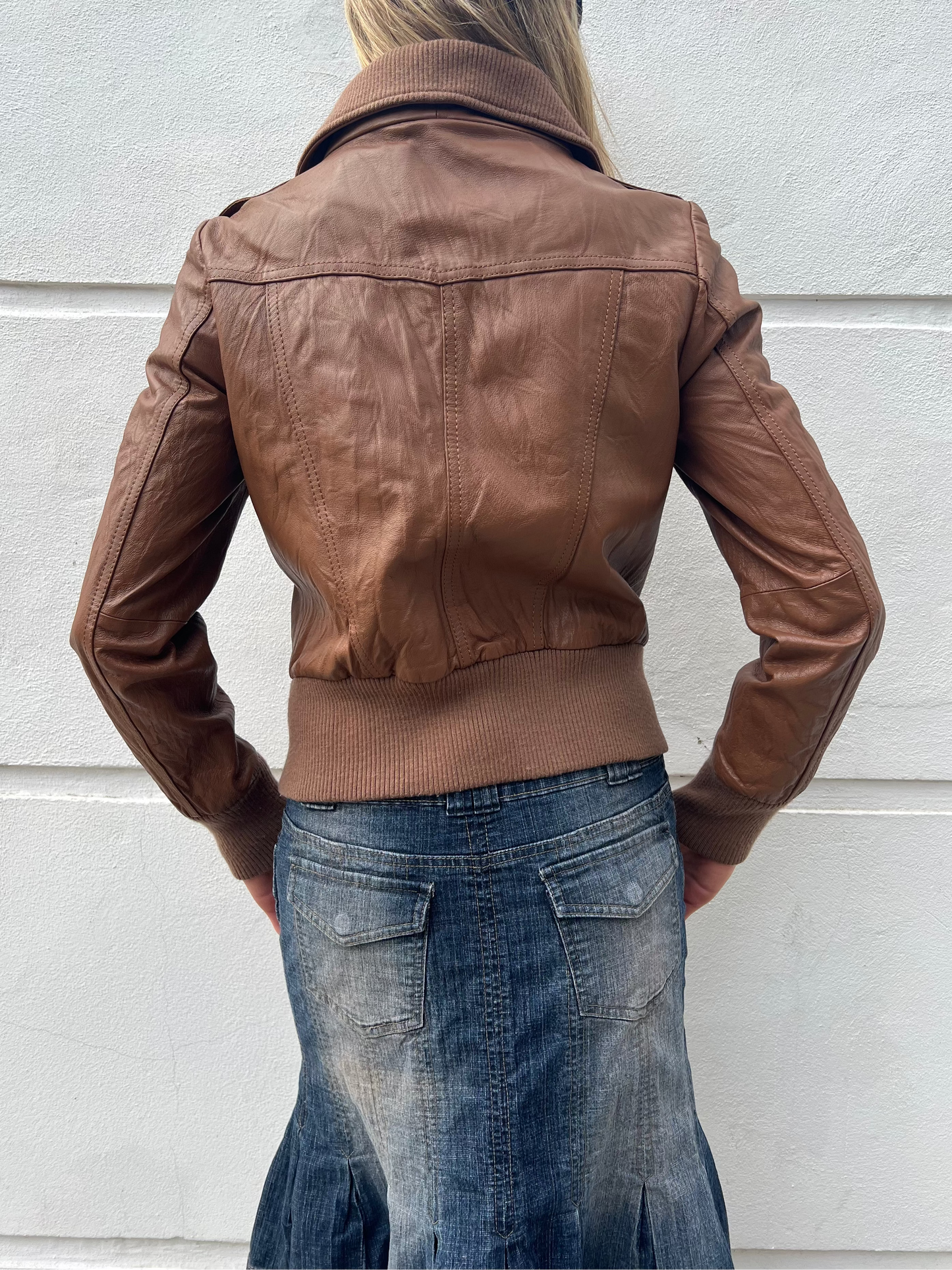 Person wearing a brown leather jacket and blue skirt against a light gray wall.