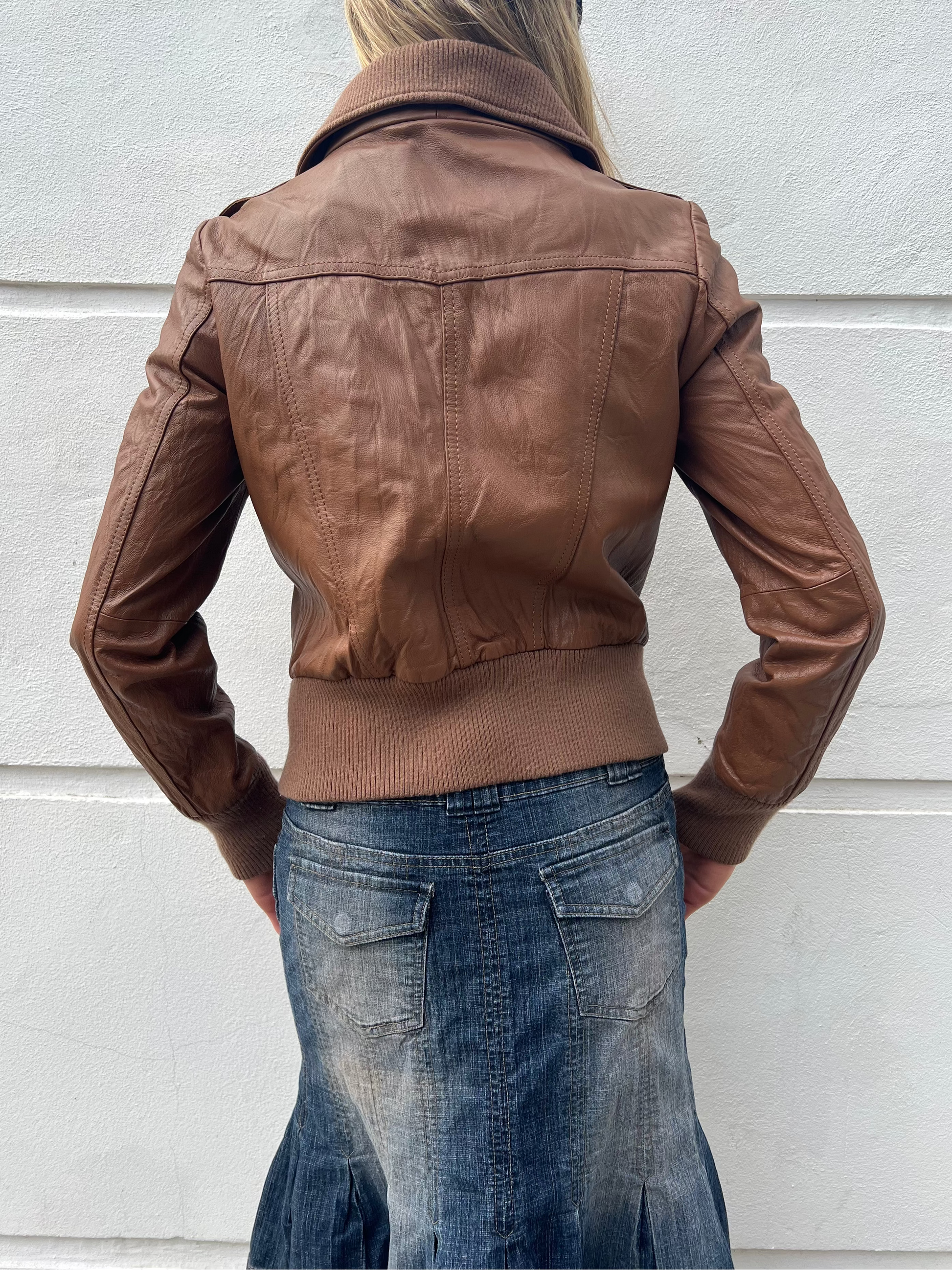 Person wearing a brown leather jacket and blue skirt against a light gray wall.