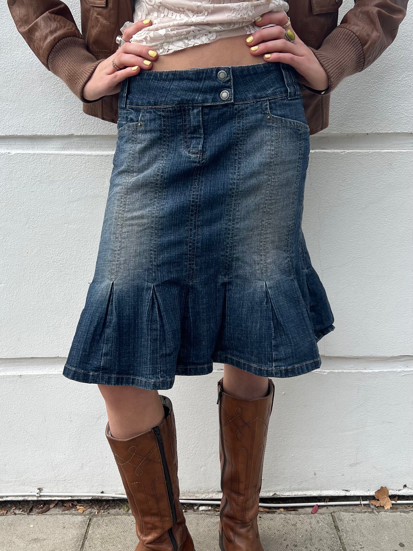 Person wearing a denim skirt and brown knee-high boots against a white wall.