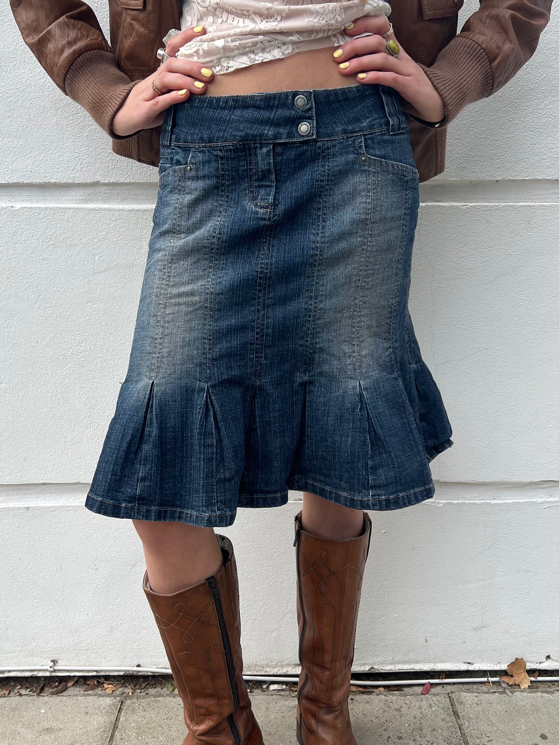 Person wearing a denim skirt and brown knee-high boots against a white wall.