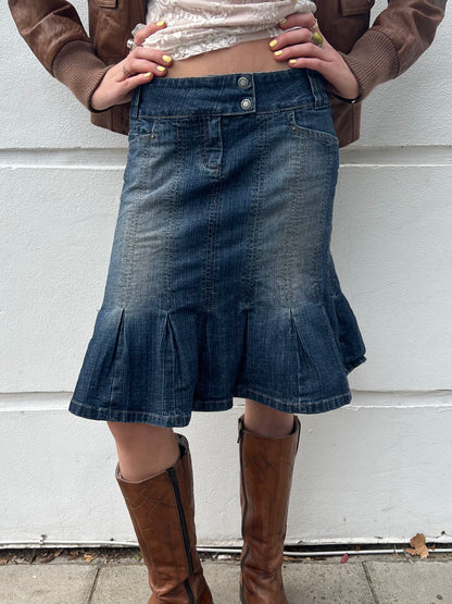 Person wearing a denim skirt and brown knee-high boots against a white wall.