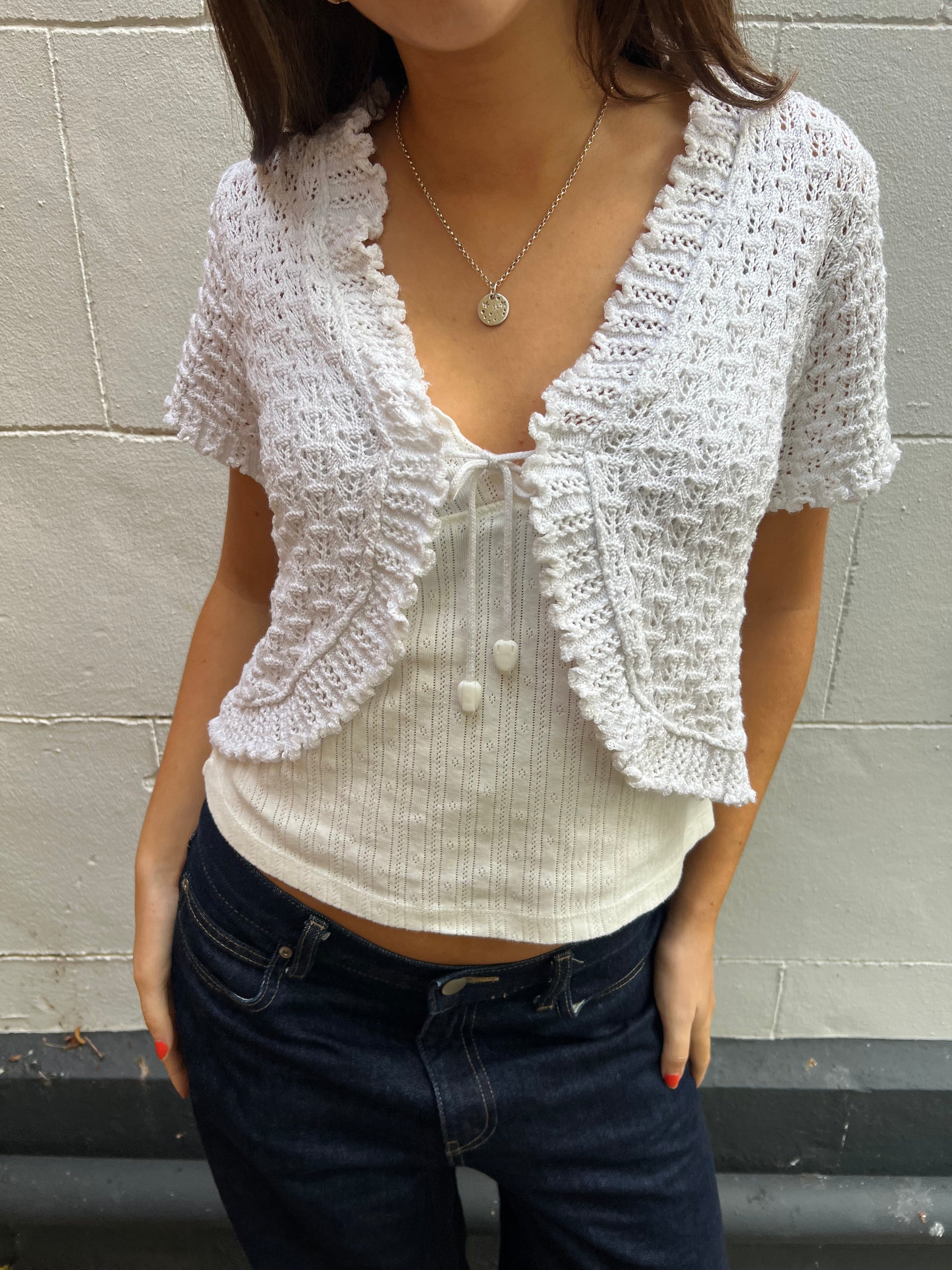 Person wearing a white textured cardigan over a white shirt with dark jeans against a plain background