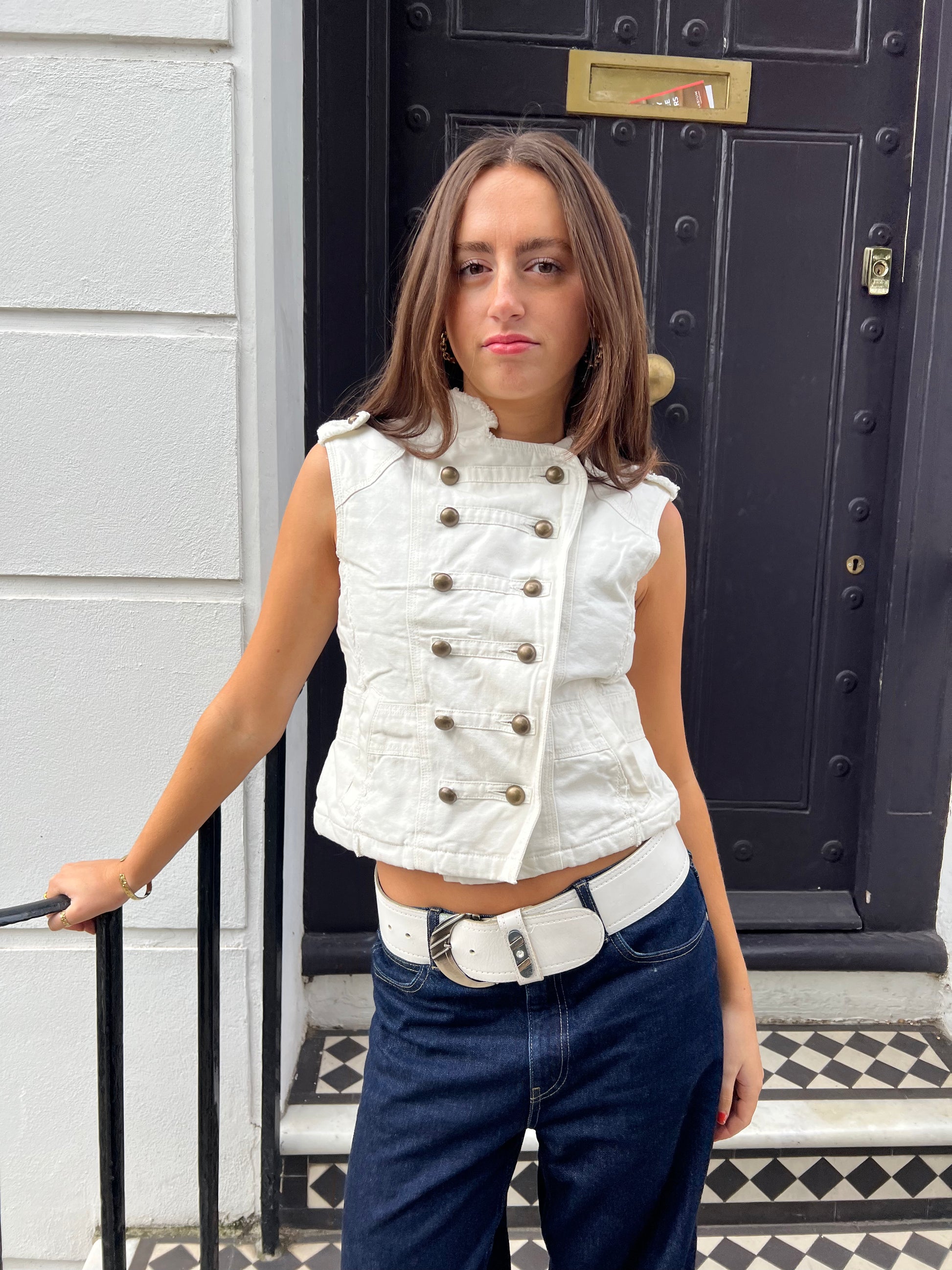 Woman wearing a white sleeveless jacket and blue jeans standing in front of a black door.